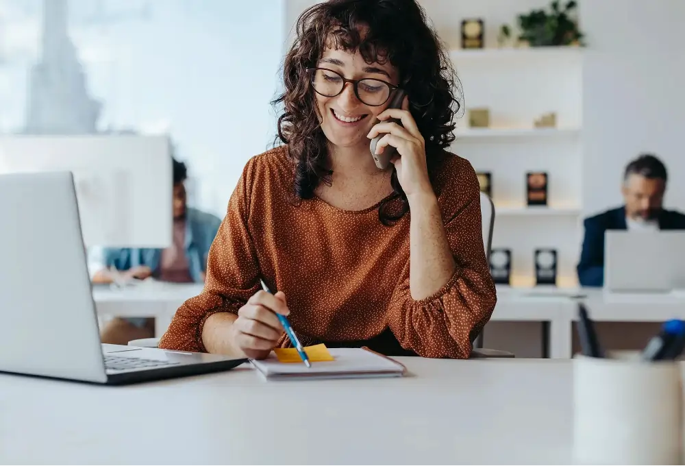 Projektmanagerin im Gespräch am Telefon in einem modernen Büro, arbeitet mit Laptop und Notizblock – symbolisch für eine Stelle im Projektmanagement für innovative Batteriesysteme.